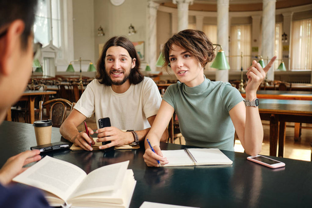 junge attraktive Gelegenheitsstudenten diskutieren in Bibliothek der Universität fröhlich über Studienprojekt - Foto, Bild
