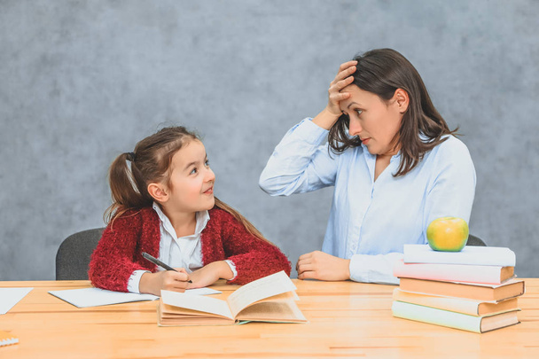 Young mother and daughter sitting on a gray background. During this, the mother watches over how a daughter performs a homework assignment. From surprise he puts his hand on his forehead. - Photo, Image