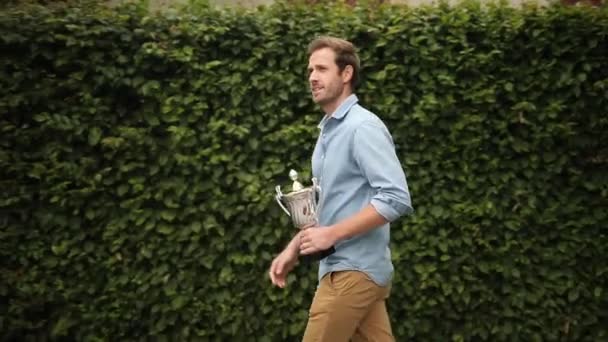 vzrušený mladý příležitostný člověk oslavuje úspěšný a přitom si drží pohár s trofejemi, skáče a na zdraví - Záběry, video