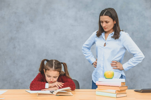 Mom and daughter on a gray background. For this, the Schoolgirl does not want to write the exercises and make a sad face looking into the book. - Foto, imagen