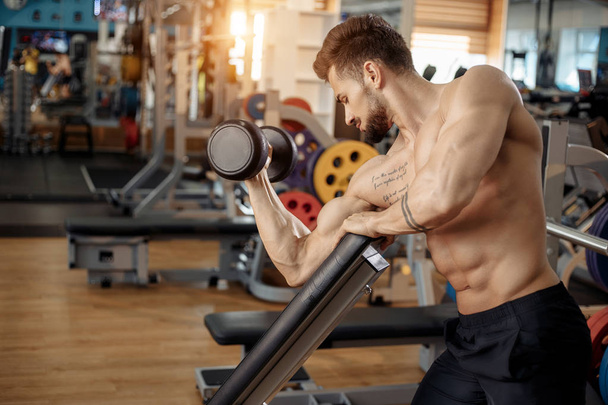 Jóképű izmos sportos fiatalember testépítő fitness modell gyakorlat bicepsz súlyzóval a padon a tornateremben. - Fotó, kép
