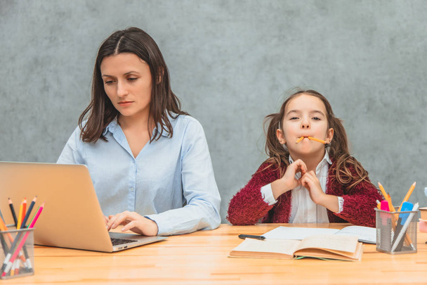Daughter and mother on a gray background. During this, the girl puts the yellow pencils in her mouth. Mom carefully reads the words during this on a laptop. - Foto, immagini