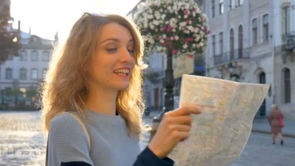 Ευτυχισμένο ενθουσιασμένο ενήλικο κορίτσι κοιτάζει τον χάρτη του χαρτιού και ψάχνοντας για κατεύθυνση νωρίς το πρωί στην αρχαία Ευρωπαϊκή πόλη σε άδειο τετράγωνο - Πλάνα, βίντεο