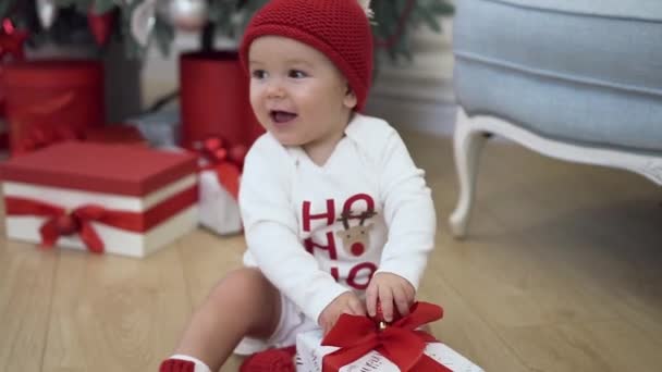 Cute smiling baby in funny new year clothes siiting near christmas tree and playing with a beautiful present - Séquence, vidéo