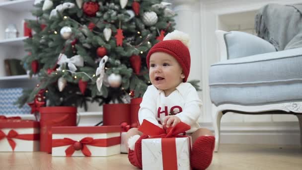 Charming toddler in amusing clothes sitting on the floor in the christmas decorated room with festive tree and playing with gift - Imágenes, Vídeo
