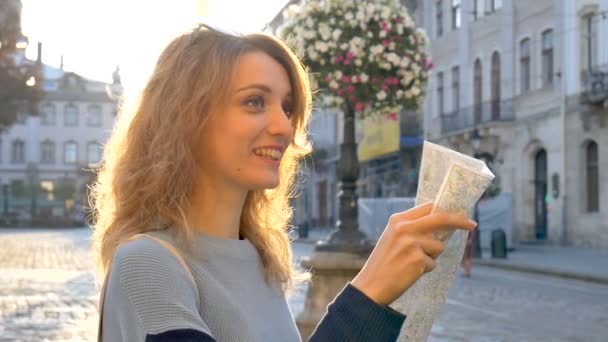Ευτυχισμένο ενθουσιασμένο ενήλικο κορίτσι κοιτάζει τον χάρτη του χαρτιού και ψάχνοντας για κατεύθυνση νωρίς το πρωί στην αρχαία Ευρωπαϊκή πόλη σε άδειο τετράγωνο - Πλάνα, βίντεο