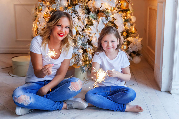 Onnellinen perhe nainen äiti ja pieni tyttö rentoutua pelaa sparkler lähellä joulukuusi jouluaattona kotona
 - Valokuva, kuva