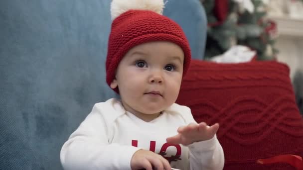 Close up of sweet toddler wearing bobble hat sitting in comfortable arm chair with pillow and playing with christmas gift - Metraje, vídeo
