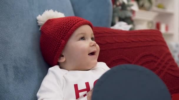 Smiling cute baby girl in bobble hat sitting in comfortable arm chair and female hand touching her nose - Materiał filmowy, wideo