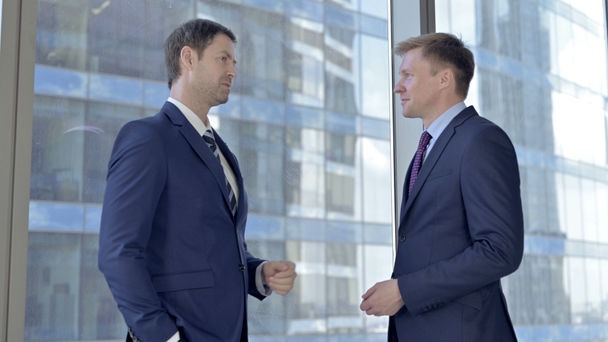 Dos hombres de negocios de mediana edad conversando y de pie contra la ventana de la oficina
 - Imágenes, Vídeo