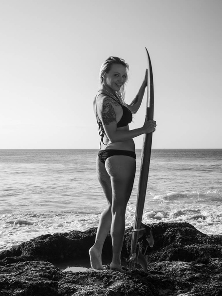 Chica surfista monocromática con tabla de surf en la playa. Mujer sexy joven con bikini negro. Chica surfista preparándose para surfear. Bali, Indonesia
 - Foto, Imagen
