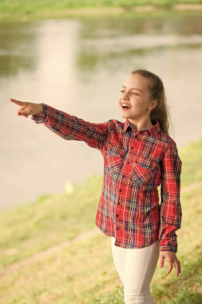 Always summer. Small girl pointing finger at river on summer landscape. Adorable child with long blond hair in casual plaid style on summer vacation. Little kid wearing fashion summer outfit - Foto, Imagen