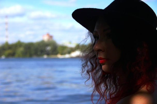 γυναίκα με κόκκινα σγουρά μαλλιά σε μαύρο καπάκι και σκούρο μίνι φόρεμα. στο ποτάμι. - Φωτογραφία, εικόνα