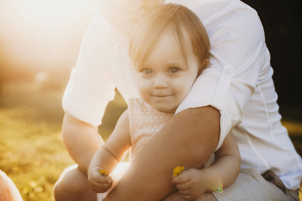 Gros plan d'une petite fille regardant la caméra sourire et tenant une fleur tout en étant embrassé par son père contre le coucher du soleil.v
 - Photo, image