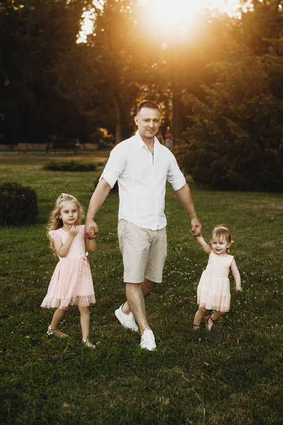 Zdjęcie młodego ojca idącego ręka w rękę z dwiema córkami przed zachodem słońca. Dwie małe dziewczynki spacerujące trzymając się za rękę z ojcem na świeżym powietrzu. - Zdjęcie, obraz