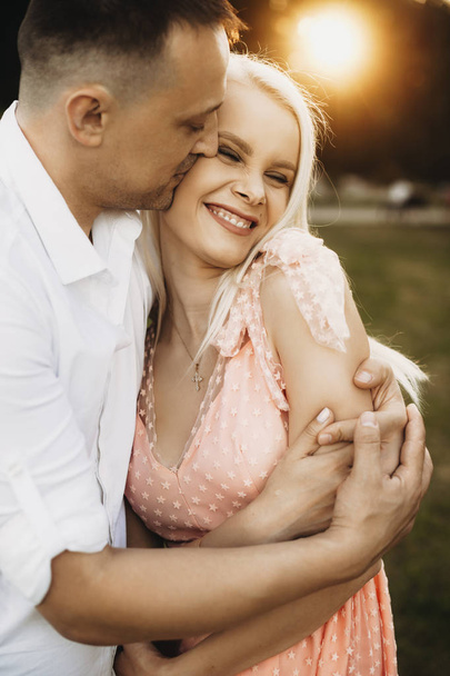 romantisches Paar, das sich draußen umarmt. Die schöne junge Frau lacht, während sie in den Armen ihres Mannes liegt. Mann mittleren Alters küsst seine Frau gegen Sonnenuntergang. - Foto, Bild