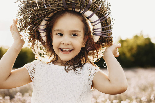 Close up portrait of a cute little girl smiling outdoor . Lovely kid wearing a wreath of flowers looking away laughing against sunset. - Photo, Image