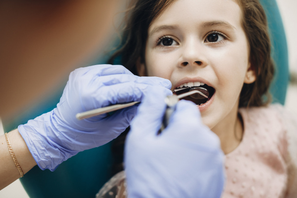 Bir pediatrik dişçinin diş muayenesi sırasında ağzı açık bir şekilde stomatoloji sandalyesinde oturan sevimli küçük bir kızın yakın çekimi.. - Fotoğraf, Görsel