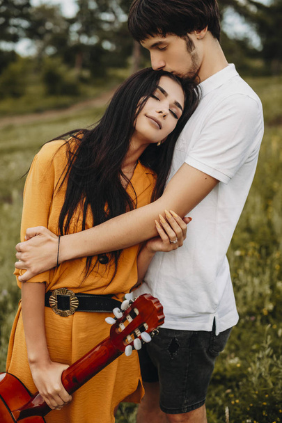 Venkovní záběr romantického páru objímajícího venkovní. Krásná mladá brunetka drží kytaru nakloněnou hlavu se zavřenýma očima na její přítel hrudníku. - Fotografie, Obrázek