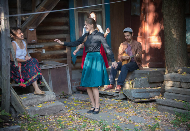Una mujer bonita con falda azul bailando junto a la música cerca de la casa de campo - un hombre jugando balalaika
 - Foto, Imagen