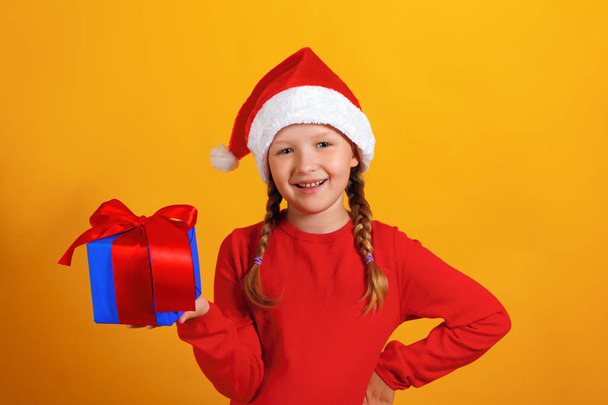 Christmas celebration. Happy little girl in santa hat holds a gift on a yellow background - Φωτογραφία, εικόνα