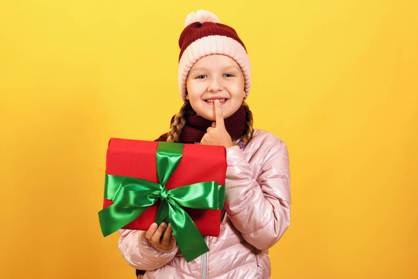 Portrait of a cute little girl in a jacket, scarf and hat on a yellow background. The child holds a box with a gift and shows a sign shh - Фото, изображение