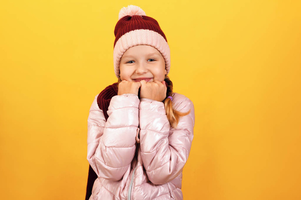 Portrait of a cute little girl in a jacket, scarf and hat on a yellow background. Autumn and winter concept - Фото, изображение