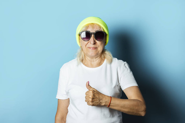 Drôle de vieille femme dans un chapeau et des lunettes, fait un geste de main sur un fond bleu. Concept cool grand-mère élégante, style moderne - Photo, image