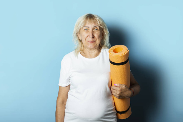 Old woman holds a yoga mat on a blue background. Yoga concept for old people, gymnastics and Pilates - Photo, Image