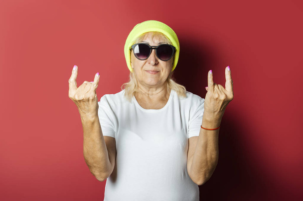 Şapkalı komik yaşlı kadın, pembe bir arka plan üzerinde el hareketi yapıyor. Konsept serin şık büyükanne, modern stil - Fotoğraf, Görsel