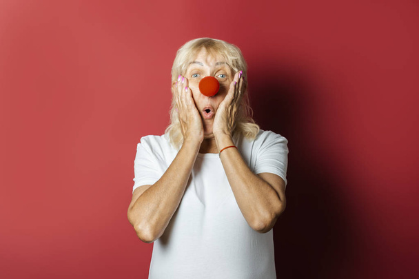 Funny old woman with a red nose of a clown on a pink background. Concept red nose day, holiday, party, clown - Photo, Image