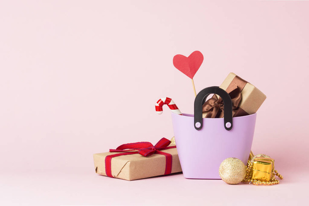 Маленький пластиковый пакет для покупок, подарочные коробки, сердце на палочке, рождественские украшения, розовый фон. Концепция предпраздничных покупок, подарки для друзей и близких, рождественская распродажа
 - Фото, изображение
