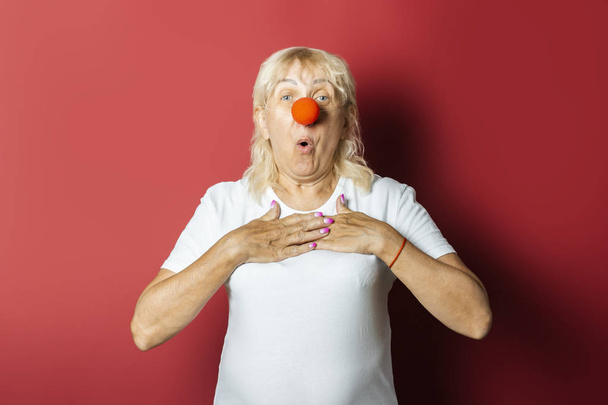 Funny old woman with a red nose of a clown on a pink background. Concept red nose day, holiday, party, clown - Photo, Image