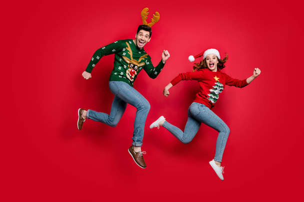 Full length photo of crazy jumping couple excited by x-mas discounts prices wear ugly ornament jumpers isolated red color background - Zdjęcie, obraz