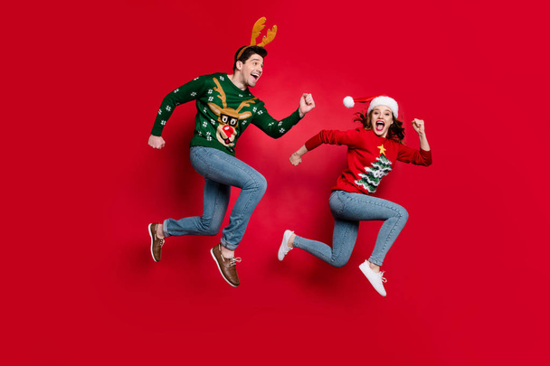 Full body photo of amazed jumping couple excited by x-mas prices hurry shopping wear ugly ornament jumpers isolated red color background - Foto, afbeelding