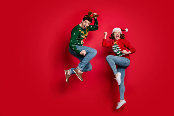 Full body photo of amazed jumping couple excited by x-mas prices wear ugly ornament jumpers and headwear isolated red color background - Photo, Image