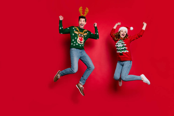 Full length photo of lady and guy jumping excited by x-mas discounts wear ugly ornament jumpers and headwear isolated red color background - Photo, Image
