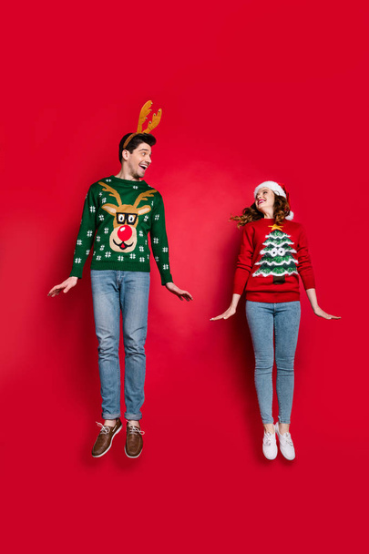 Vertical full size photo of lady and guy jumping high excited by x-mas discounts wear knitted pullovers and headwear isolated red color background - 写真・画像