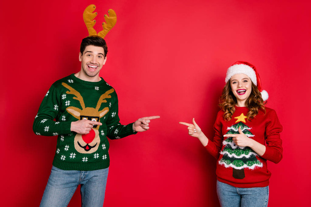 Photo of funny lady and guy directing fingers empty space advising good choice wear knitted ugly ornament pullovers isolated red color background - Foto, Bild