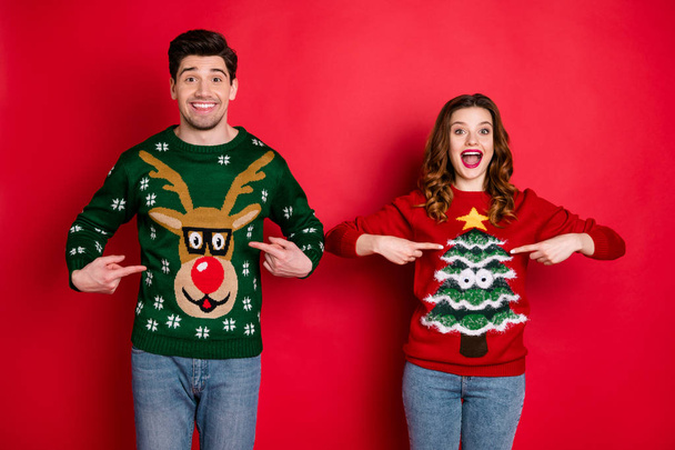 Midnight presents. Portrait of two brunet hair lovers people scream wow omg point indexf inger his her reindeer christmas tree pattern pullovers wear jeans isolated over red color background - Φωτογραφία, εικόνα