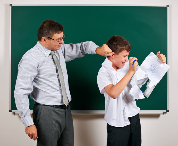 Portrait of a teacher catch the ear funny schoolboy with low discipline. Pupil very emotional, having fun and very happy, posing at blackboard background - back to school and education concept - Fotoğraf, Görsel