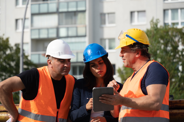 Trois travailleurs sur le chantier portant un gilet de sécurité, un casque et des lunettes fournissant des instructions à l'aide d'une tablette numérique
 - Photo, image