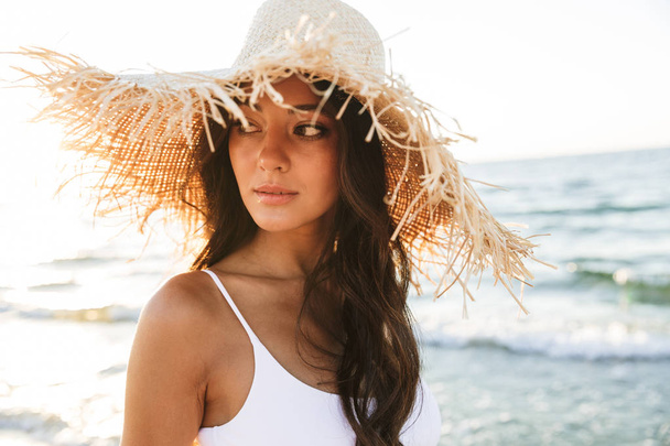 Photo of thoughtful brunette woman in swimsuit and straw hat wal - Фото, изображение