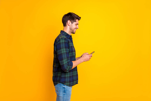 Profile photo of blogger guy holding telephone in hands checking instagram followers wear casual plaid shirt isolated yellow color background - Foto, Imagen