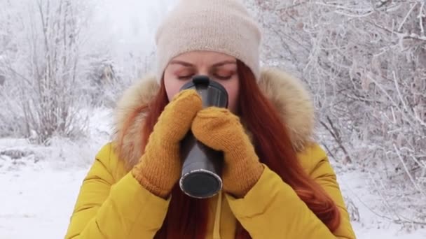Joven pelirroja con un cálido sombrero de punto y una chaqueta de invierno amarilla bebe una bebida caliente de una taza metálica sobre el fondo de un paisaje invernal
. - Metraje, vídeo