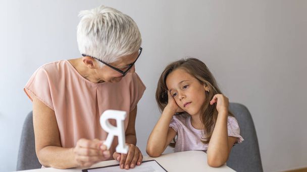 Little girl with teacher on private lesson. A child with development problems with a professional speech therapist during a meeting. Tutor holding a letter 'R'. - Foto, afbeelding