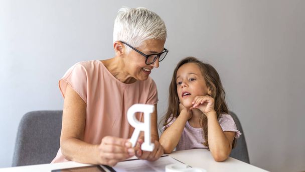 Girl visiting speech therapist. Mature female therapist helping girl in speech therapy exercise. Cute little girl at speech therapist office. Mature woman teacher and little girl on private lesson - Foto, afbeelding