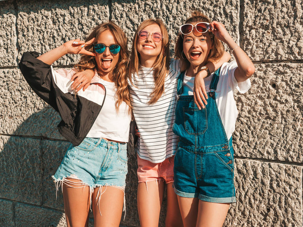 Portrait of three young beautiful smiling hipster girls in trendy summer clothes. Sexy carefree women posing near wall in the street.Positive models having fun in sunglasses - 写真・画像