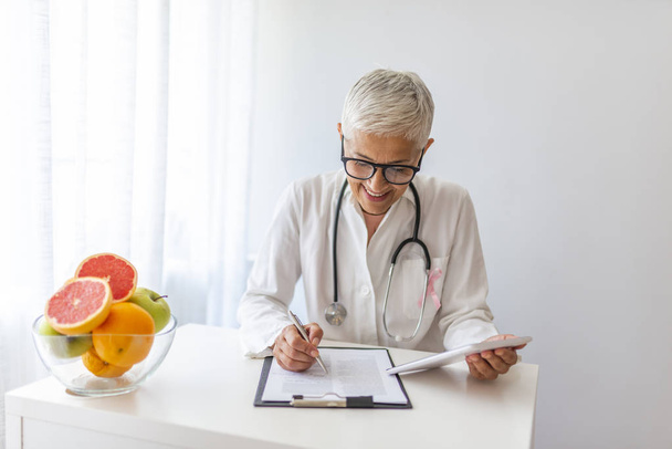Female dietician showing vegetables and fruit. Doctor writing on paperwork. Doctor nutritionist writing case history in the office. Senior woman dietitian prescribing recipe - Foto, Imagem