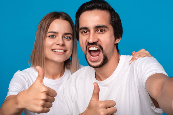 Happy couple of a young blond woman and brunet bearded man with mustaches in white t-shirts and blue jeans making selfie holding thumbs up isolated over blue background. Concept of an ideal couple. - Photo, image